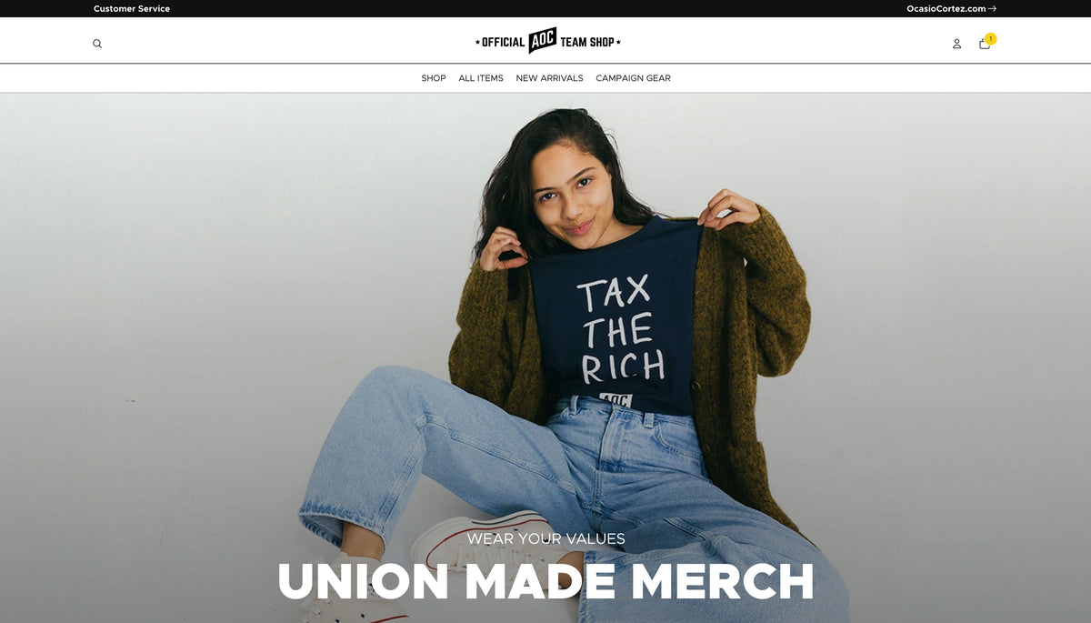 A woman sitting against a light gray wall wearing a navy graphic T-shirt that reads 'TAX THE RICH' in white block letters, layered under a brown cardigan and light blue jeans. The image appears to be a promotional or website-style crop with large text at the bottom reading 'UNION MADE MERCH'.
